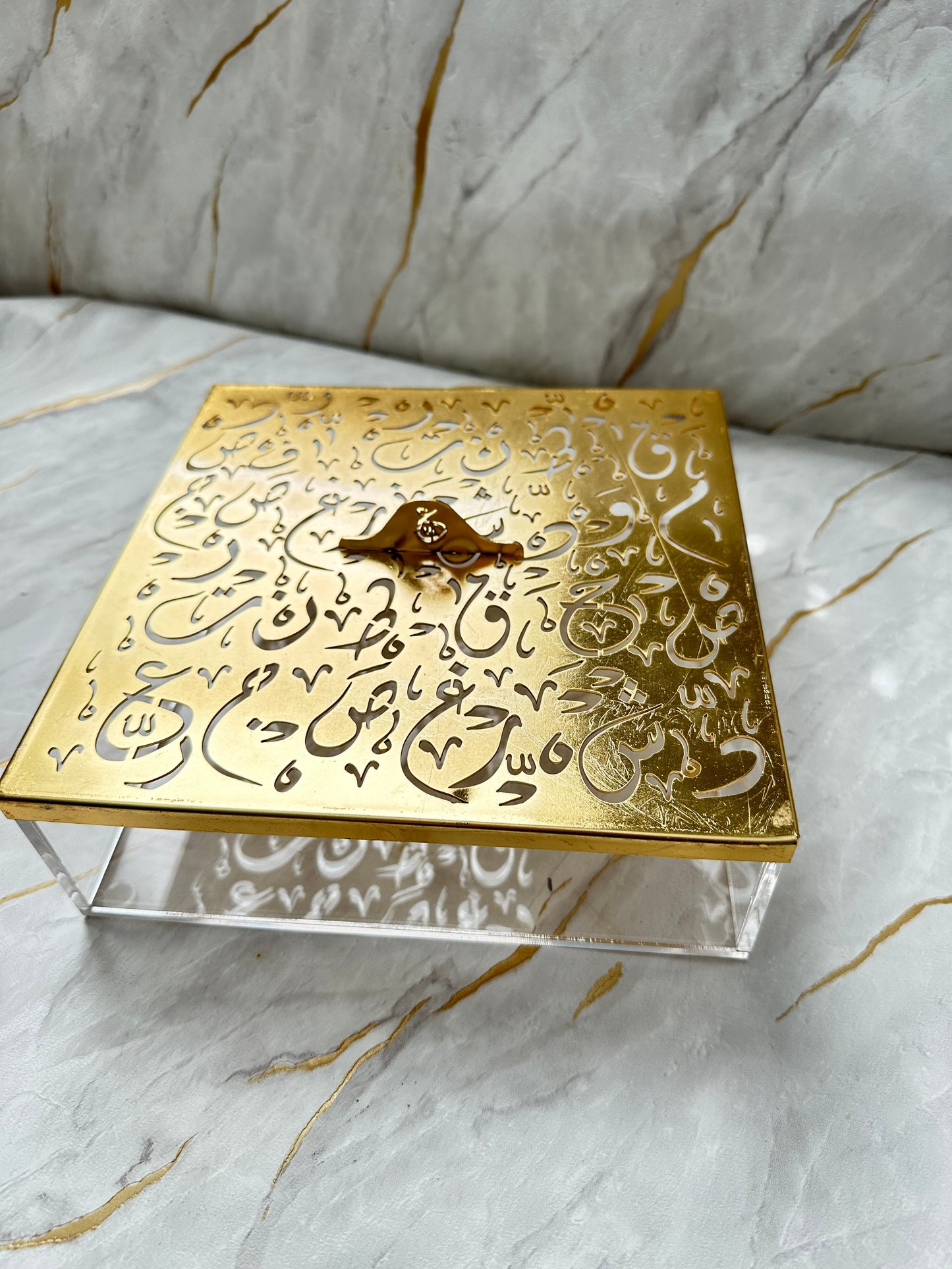 Discover elegance with our gold-tone decorative box featuring intricate Arabic calligraphy. This stylish piece not only offers ample storage but also serves as a sophisticated addition to any space. The polished gold lid complements the transparent acrylic base, striking a balance between modern aesthetics and cultural heritage. Whether as a gift or a personal indulgence, this spacious box is a statement of refined taste and artistic appreciation.