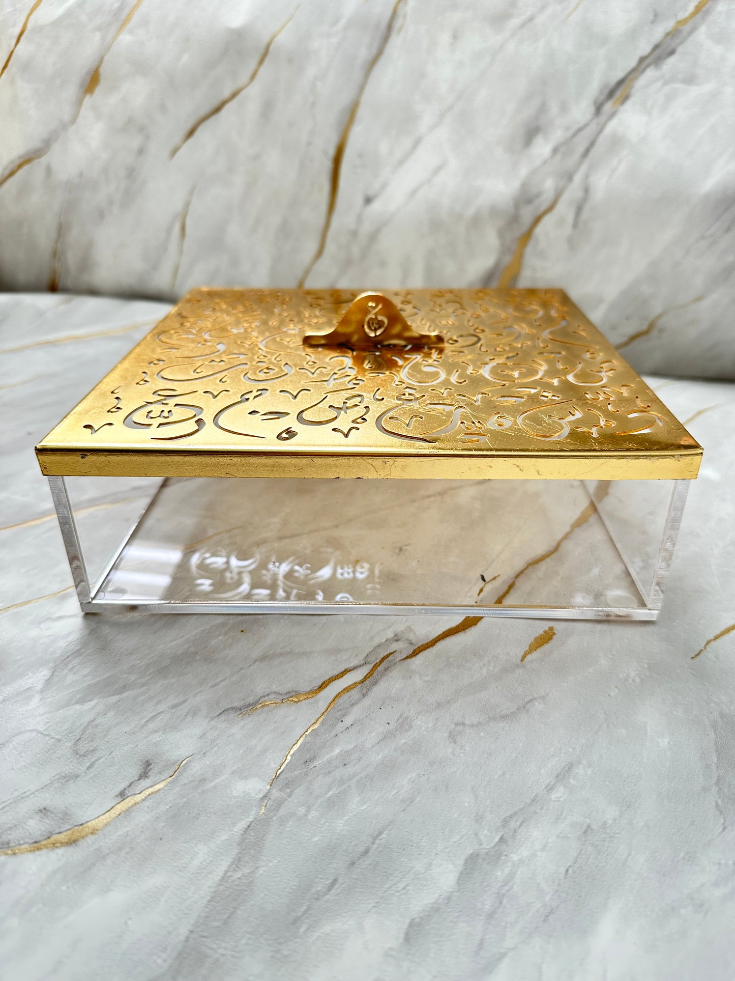 Add a touch of sophistication to your decor with our gold-tone decorative box, adorned with intricate Arabic calligraphy. This versatile piece doubles as a practical storage solution and a stylish accent for any room. The polished gold lid contrasts beautifully with the transparent acrylic base, creating a sleek and modern aesthetic. Whether as a gift or a personal addition to your collection, this spacious box embodies elegance and cultural richness.